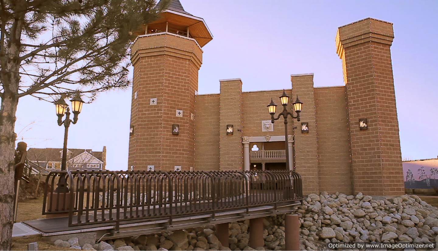 Castle Park's stone castle building at dusk, with two tall brick towers, string lights along the crenellated walls, and an iron-railed bridge-style walkway leading to the entrance